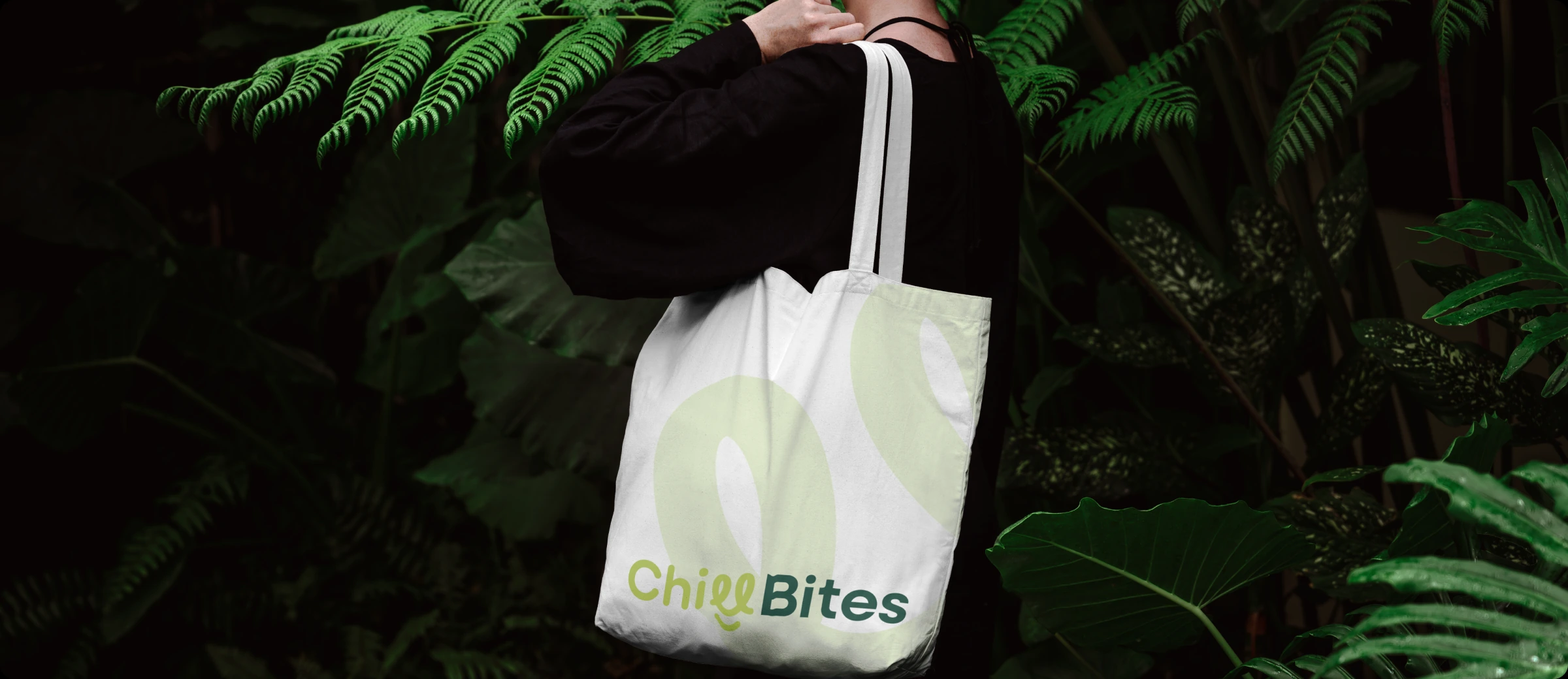 A person carrying a white ChillBites tote bag among tropical leaves